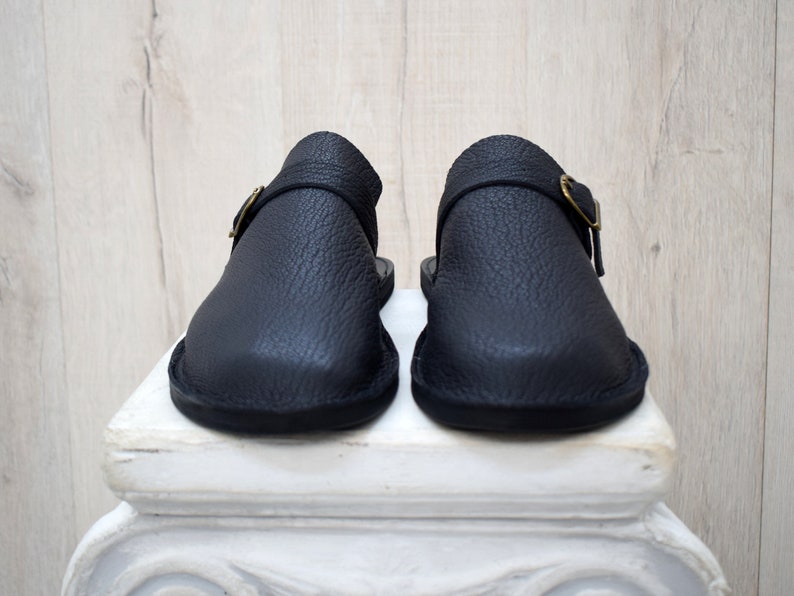 May include: A pair of black leather clogs with a textured finish. Each clog features a strap with a gold-colored buckle. The shoes are displayed on a white pedestal against a light wood background.