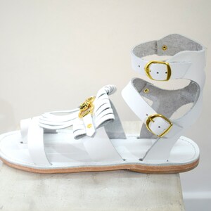 May include: White leather gladiator sandals with gold-tone buckles and accents. The sandals feature a toe loop, ankle straps, and decorative fringe. The sole is a light brown color.