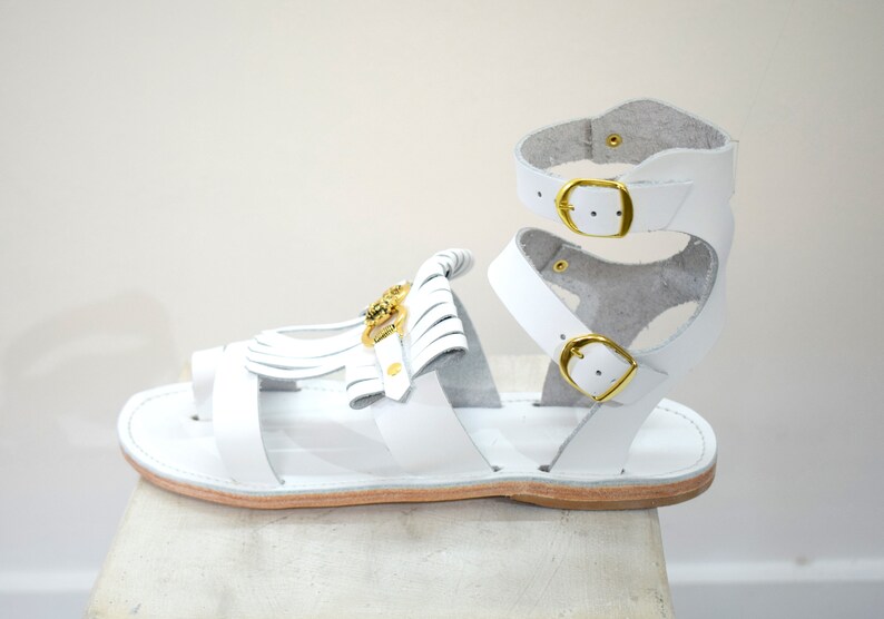 May include: White leather gladiator sandals with gold-tone buckles and accents. The sandals feature a toe loop, ankle straps, and decorative fringe. The sole is a light brown color.