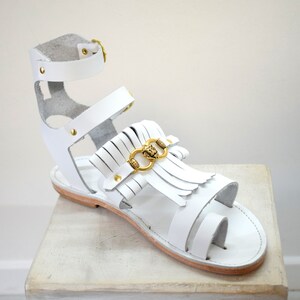 May include: White leather gladiator sandal with gold-colored accents. The sandal features a toe loop, multiple straps, and a fringe detail with a gold-colored lion head emblem. The sandal is displayed on a light-colored surface.
