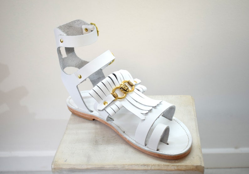 May include: White leather gladiator sandal with gold-colored accents. The sandal features a toe loop, multiple straps, and a fringe detail with a gold-colored lion head emblem. The sandal is displayed on a light-colored surface.