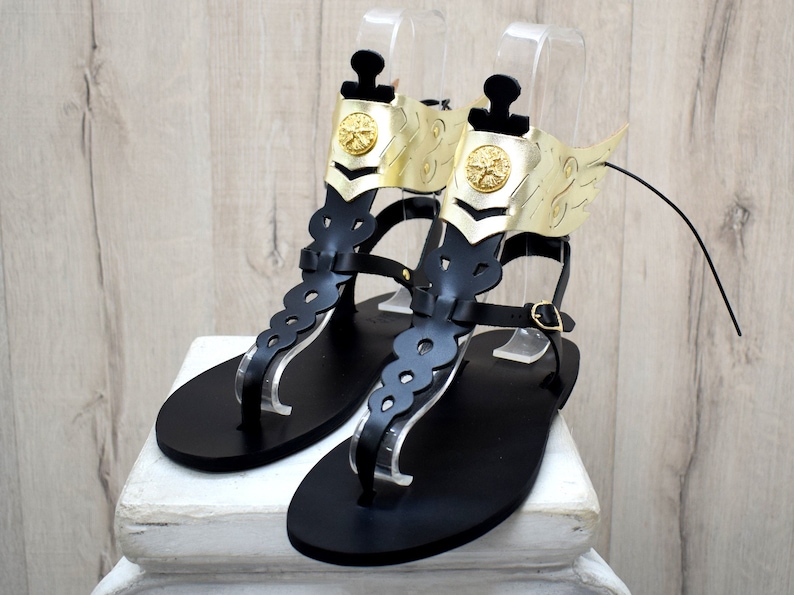 May include: A pair of black leather sandals featuring a tall, cutout vertical strap and a gold-colored metallic ankle cuff. The cuff is adorned with a circular emblem and a stylized wing extension on each side, secured with a thin black buckle strap.
