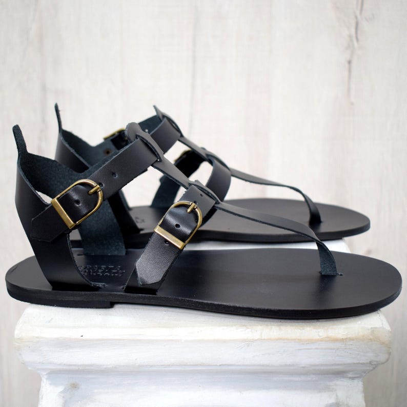 Puede incluir: Un par de sandalias de cuero negro con m&uacute;ltiples correas y hebillas doradas. Las sandalias tienen una suela plana y una correa para el dedo gordo del pie tipo tanga. Las sandalias se exhiben sobre una superficie blanca.