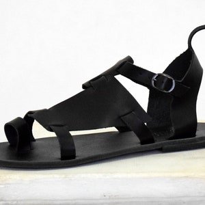 K&ouml;nnte beinhalten: Schwarze Ledersandalen mit Schnallenriemen und Zehenschlaufe. Die Sandalen haben eine flache Sohle und sind aus weichem, geschmeidigem Leder gefertigt.