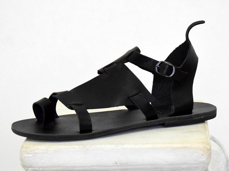 K&ouml;nnte beinhalten: Schwarze Ledersandalen mit Schnallenriemen und Zehenschlaufe. Die Sandalen haben eine flache Sohle und sind aus weichem, geschmeidigem Leder gefertigt.