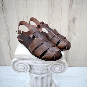 May include: Brown leather sandals with a caged design. The sandals have a strap around the ankle and a buckle closure. The sandals are displayed on a white pedestal with a decorative design.