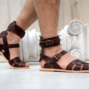May include: Brown leather sandals with multiple straps and buckle closures. The sandals have a light-colored sole and are designed in a gladiator style. The image shows the sandals on a person's feet.