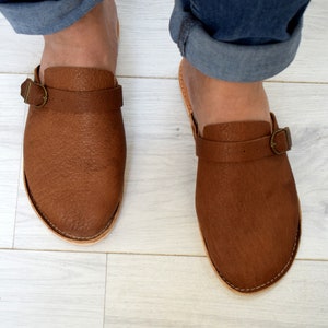 May include: A pair of brown leather slip-on clogs with a buckle strap. The clogs have a rounded toe and a stitched edge.