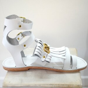 May include: White leather sandal with gold-tone hardware. The sandal features multiple straps, including an ankle strap with a buckle closure and a decorative fringe detail. The sole is a light brown color.
