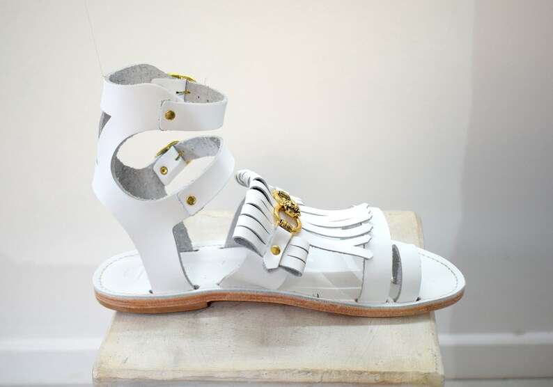 May include: White leather sandal with gold-tone hardware. The sandal features multiple straps, including an ankle strap with a buckle closure and a decorative fringe detail. The sole is a light brown color.