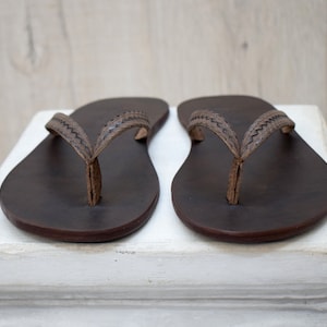 Op de afbeelding: Een paar donkerbruine leren teenslippers met gestikte bandjes. De sandalen worden gepresenteerd op een wit oppervlak tegen een lichte houten achtergrond. Deze teenslippers zijn een klassieke stijl, geschikt voor vrijetijdskleding.