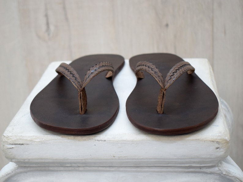 Op de afbeelding: Een paar donkerbruine leren teenslippers met gestikte bandjes. De sandalen worden gepresenteerd op een wit oppervlak tegen een lichte houten achtergrond. Deze teenslippers zijn een klassieke stijl, geschikt voor vrijetijdskleding.