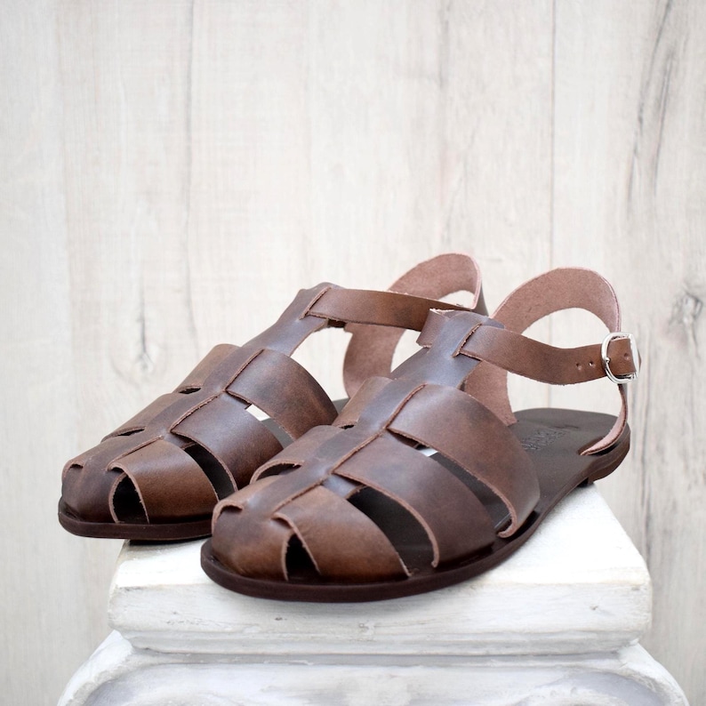 May include: A pair of brown leather sandals with a cage-like design. The sandals have a strap that buckles on the side and are displayed on a white surface against a light wood background. The sandals are a medium brown color.