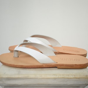 Peut inclure: Une paire de tongs en cuir blanc avec une semelle marron clair. Les sandales ont un design simple avec une bride en lani&egrave;re et une semelle plate. Les sandales sont expos&eacute;es sur une surface blanche.
