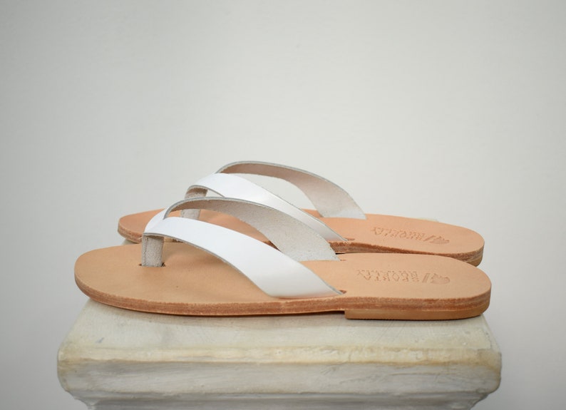 Peut inclure: Une paire de tongs en cuir blanc avec une semelle marron clair. Les sandales ont un design simple avec une bride en lani&egrave;re et une semelle plate. Les sandales sont expos&eacute;es sur une surface blanche.