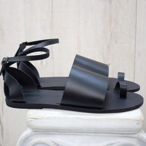 May include: A pair of black leather sandals with a minimalist design. The sandals feature a toe loop, ankle strap with buckle closure, and wide straps across the foot. The sandals are displayed on a white surface against a light wood background.