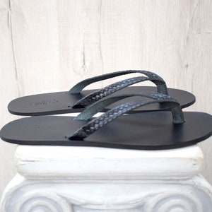 Op de afbeelding: Een paar zwarte leren teenslippers met gevlochten bandjes. De sandalen worden getoond op een witte decoratieve zuil tegen een lichte houtnerf achtergrond. De teenslippers hebben een minimalistisch ontwerp.