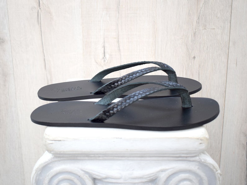 Op de afbeelding: Een paar zwarte leren teenslippers met gevlochten bandjes. De sandalen worden getoond op een witte decoratieve zuil tegen een lichte houtnerf achtergrond. De teenslippers hebben een minimalistisch ontwerp.