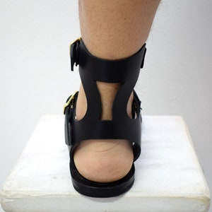 May include: Black leather gladiator sandals with gold-colored buckles. The sandals feature a cutout design on the ankle strap and a flat sole. The image shows the back of the sandal and part of the leg.
