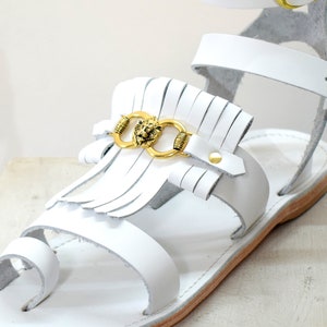 May include: White leather sandals with a gold lion head accent and fringe detail. The sandals have a toe loop and ankle strap with a gold buckle. The sole is a light brown color.