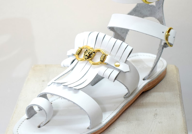 May include: White leather sandals with a gold lion head accent and fringe detail. The sandals have a toe loop and ankle strap with a gold buckle. The sole is a light brown color.