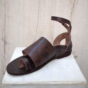 May include: Brown leather sandal with a toe loop and ankle strap. The sandal features a wide strap across the top of the foot. The sole is a dark brown color, and the sandal appears to be handmade.