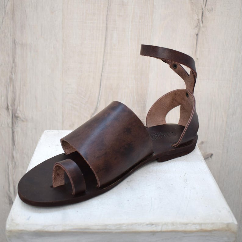 May include: Brown leather sandal with a toe loop and ankle strap. The sandal features a wide strap across the top of the foot. The sole is a dark brown color, and the sandal appears to be handmade.