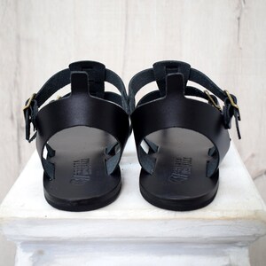 Puede incluir: Un par de sandalias de cuero negro con m&uacute;ltiples correas y hebillas doradas. Las sandalias se muestran desde la parte trasera, mostrando su dise&ntilde;o y construcci&oacute;n. Las sandalias est&aacute;n sobre una superficie blanca.