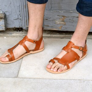 K&ouml;nnte beinhalten: Ein Paar braune Ledersandalen mit mehreren Riemen und einem Schnallenverschluss. Die Sandalen werden von einer Person getragen.
