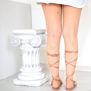 May include: A pair of tan leather gladiator sandals featuring long, thin laces that wrap around the lower legs and tie in a knot at the back of the calf. The sandals have a flat sole and a minimalist design, showcasing the light brown straps.