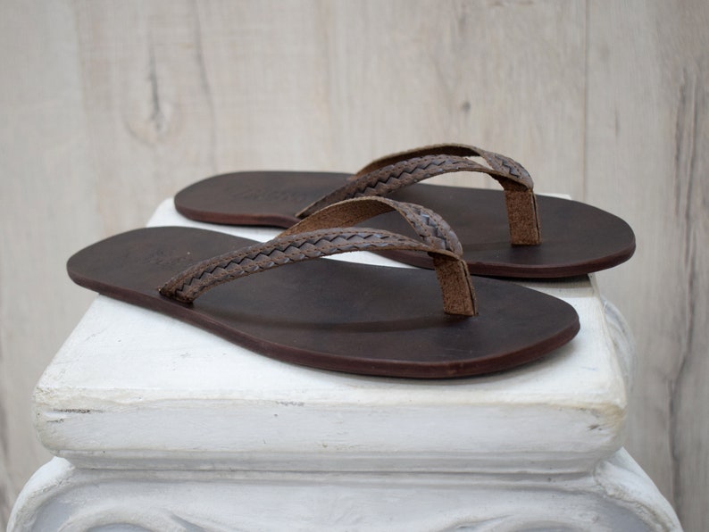 Op de afbeelding: Een paar bruine leren slippers. De sandalen hebben een donkerbruine zool en een gevlochten band. De teenslipper is lichtbruin van kleur. De slippers worden op een wit oppervlak gepresenteerd.