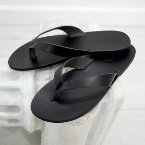 Peut inclure: Une paire de tongs en cuir noir. Les sandales ont un design simple avec une bride en lani&egrave;re et une semelle plate. Le cuir semble lisse et les sandales sont pr&eacute;sent&eacute;es sur une surface blanche.
