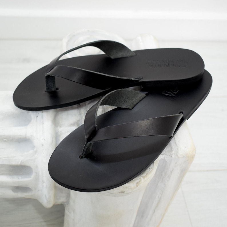 Peut inclure: Une paire de tongs en cuir noir. Les sandales ont un design simple avec une bride en lani&egrave;re et une semelle plate. Le cuir semble lisse et les sandales sont pr&eacute;sent&eacute;es sur une surface blanche.