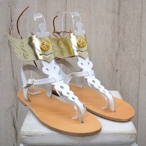 May include: A pair of white leather sandals featuring gold-colored metallic wing attachments at the ankles. Each sandal includes a central vertical strap with decorative cutouts, a gold circular emblem, and a thin brown leather lace for closure.