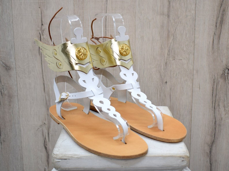 May include: A pair of white leather sandals featuring gold-colored metallic wing attachments at the ankles. Each sandal includes a central vertical strap with decorative cutouts, a gold circular emblem, and a thin brown leather lace for closure.