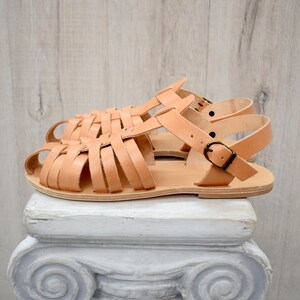 May include: A pair of light brown leather sandals with a woven strap design. The sandals have adjustable ankle straps with metal buckles. The sandals are displayed on a white pedestal against a light wood background.