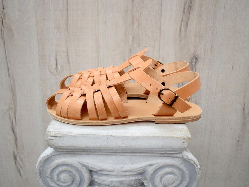 May include: A pair of light brown leather sandals with a woven strap design. The sandals have adjustable ankle straps with metal buckles. The sandals are displayed on a white pedestal against a light wood background.