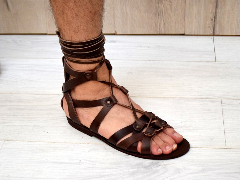 Puede incluir: Una sandalia de gladiador de cuero marr&oacute;n oscuro con m&uacute;ltiples correas horizontales sobre el pie y un cord&oacute;n largo que se envuelve alrededor del tobillo. La sandalia tiene una suela plana y est&aacute; asegurada con peque&ntilde;os remaches met&aacute;licos.