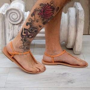 May include: Tan leather sandals with orange straps and a buckle closure. The sandals have a simple T-strap design and a flat sole. The image shows the sandals on a person's feet, showcasing the minimalist style.