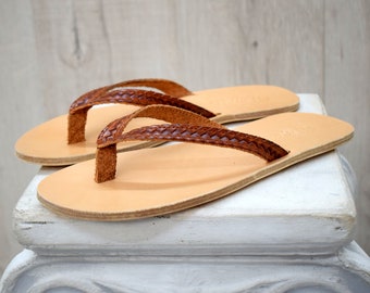 May include: A pair of flat sandals featuring light tan, smooth leather footbeds and dark brown, braided leather straps. The thong-style straps connect to the sole between the first and second toes, creating a simple, open design for these footwear items.