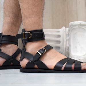 May include: Black leather sandals with multiple straps and gold-colored buckles. The sandals have a flat sole and a high ankle strap. The design is reminiscent of ancient Roman footwear. The sandals are worn by a person.