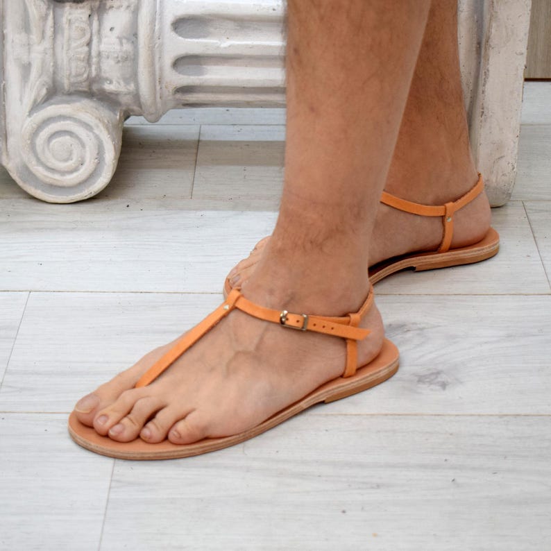 May include: Orange leather sandals with a T-strap design and adjustable ankle straps. The sandals have a flat sole and a small buckle closure. The footwear is shown on a person's feet.