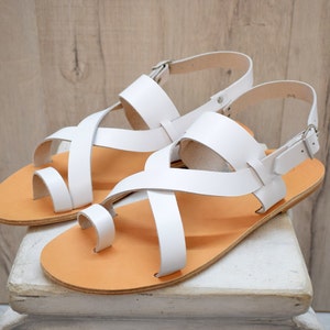 Peut inclure: Une paire de sandales blanches avec un design à boucle d'orteil et de larges brides croisées sur le dessus du pied. Les sandales possèdent une semelle plate beige et une bride de cheville réglable avec une boucle en métal argenté.