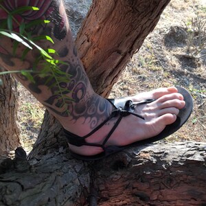 May include: A person's foot wearing black leather sandals with a thick sole and laces that wrap around the ankle. The sandals are worn on a tree branch.