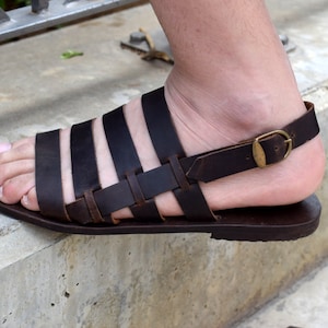 May include: Brown leather sandals with a buckle strap and multiple straps across the foot.