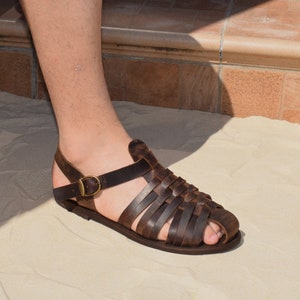 May include: Brown leather sandal with a woven design across the foot and a strap around the ankle. The sandal features a buckle closure and is designed for comfort and style. The leather has a distressed look, adding to its rustic appeal.