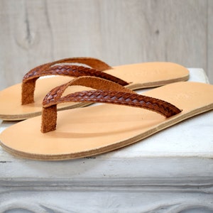 May include: A pair of brown leather thong sandals with a braided strap. The sandals are on a white pedestal.