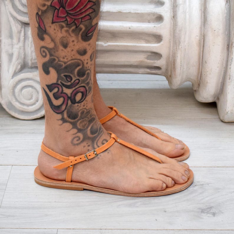 May include: Orange leather sandals with a T-strap design. The sandals have a buckle closure and a flat sole. The leather is a light orange color. The sandals are designed for warm weather and casual wear.