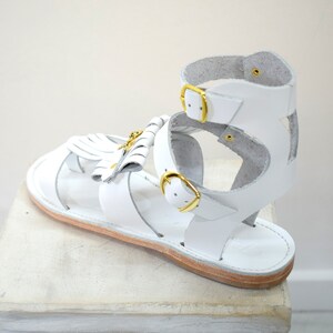 May include: White leather sandal with gold-colored buckles and a decorative tassel. The sandal features a strappy design with an ankle strap and a flat sole. The sandal is displayed on a white surface.
