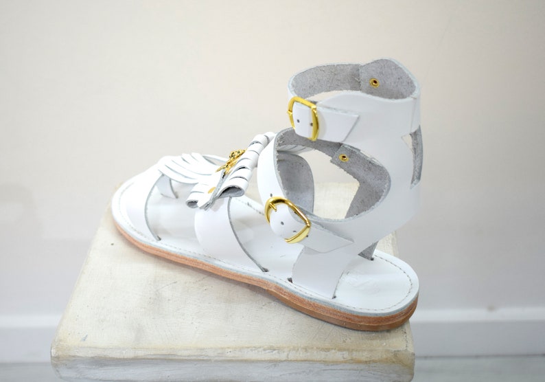 May include: White leather sandal with gold-colored buckles and a decorative tassel. The sandal features a strappy design with an ankle strap and a flat sole. The sandal is displayed on a white surface.